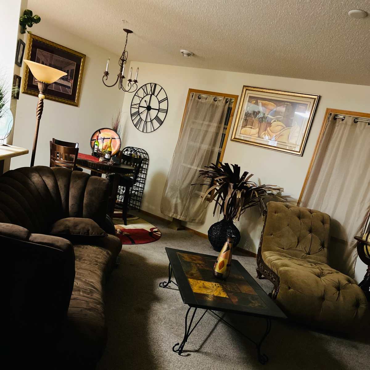 A living room with a brown sofa, an armchair, a glass coffee table, a dining area with a clock on the wall, and decor items including a lamp and plants.