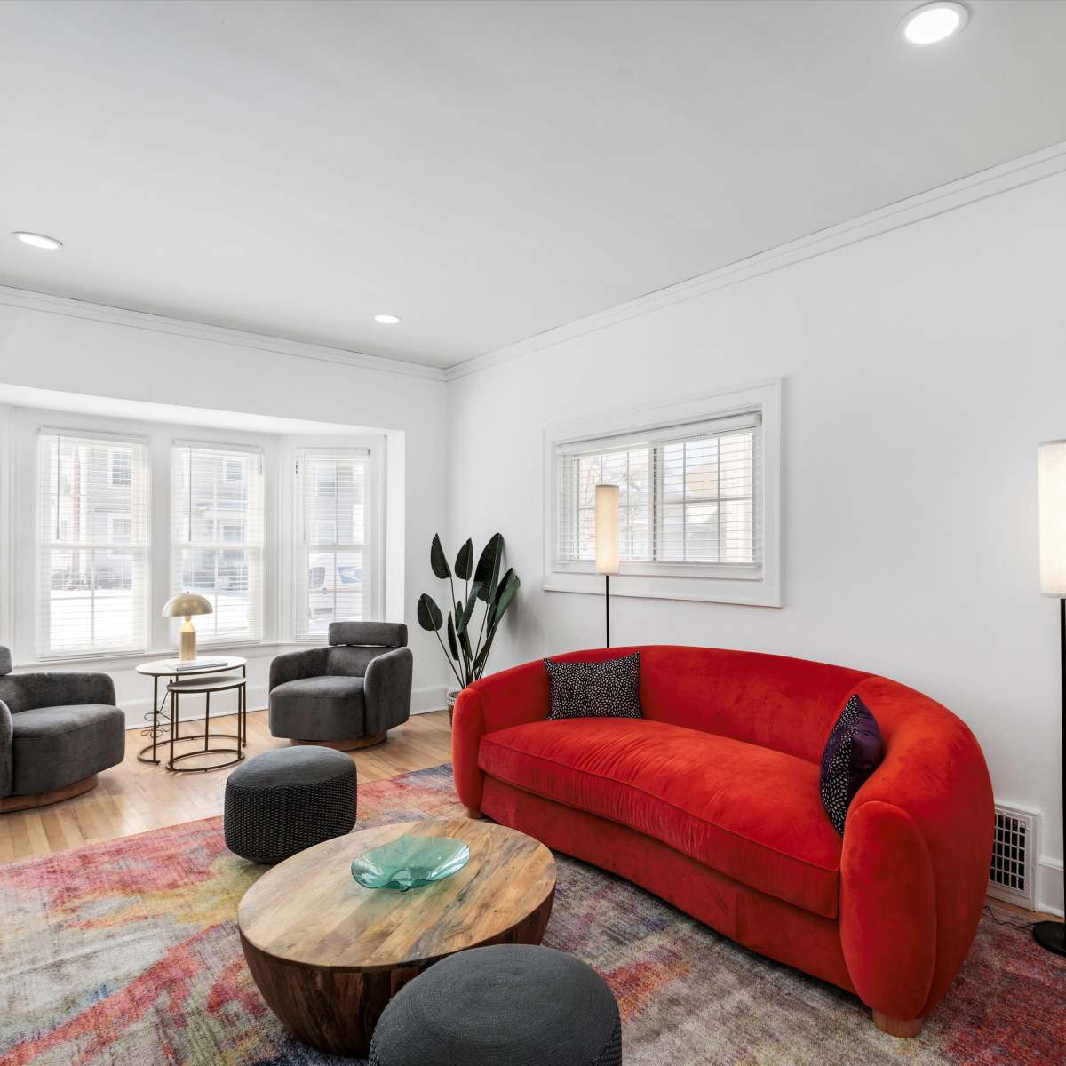 The living room features a red sofa, two gray armchairs, a round wooden coffee table, and natural light flowing through bay windows.
