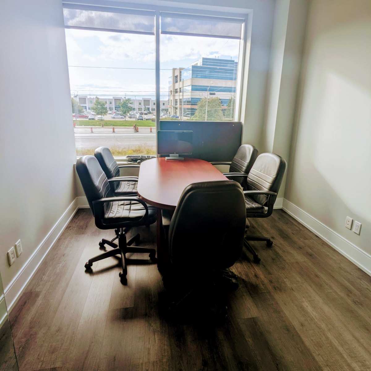 A small conference room with a round table and four black chairs near a large window, overlooking a street and buildings outside.