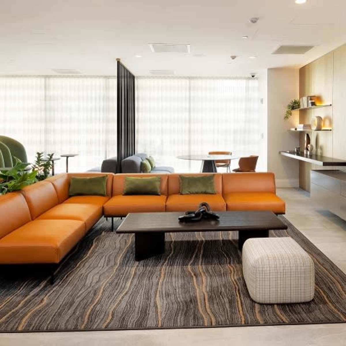 A modern lounge area with a large orange sectional sofa, a black coffee table, and a neutral-patterned rug, surrounded by large windows and contemporary decor.