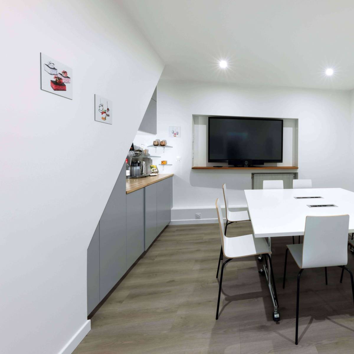 A modern conference room features a large white table surrounded by eight chairs, a wall-mounted television, and framed artwork on the walls.