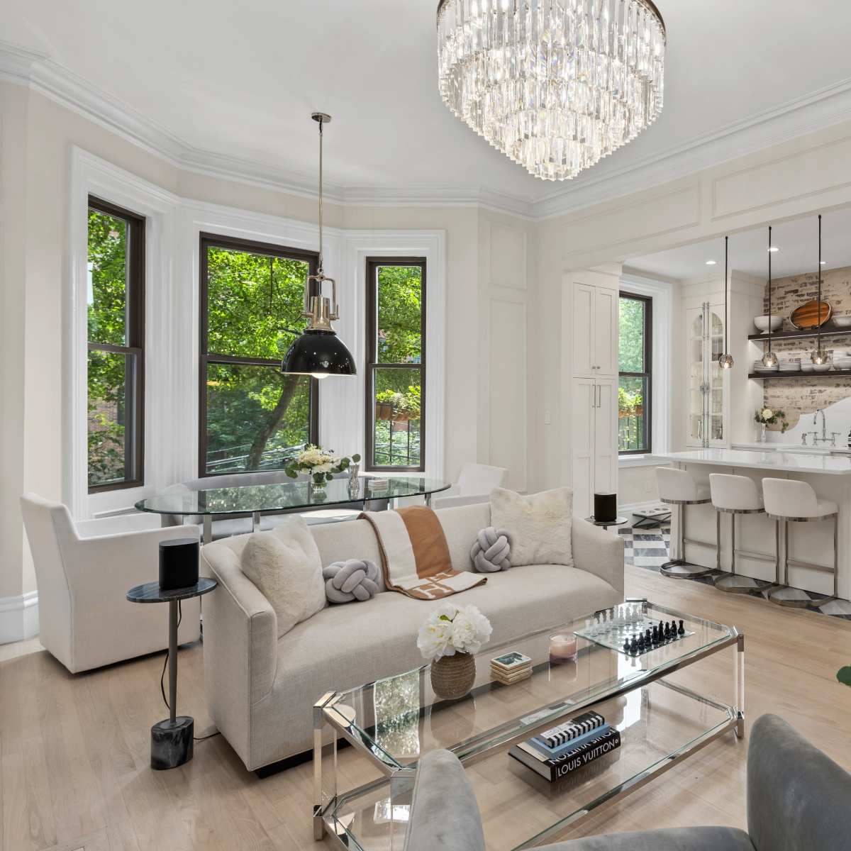 A modern living room featuring a light-colored sofa and chairs, a glass coffee table, and a kitchen visible in the background with open shelves and stylish decor.