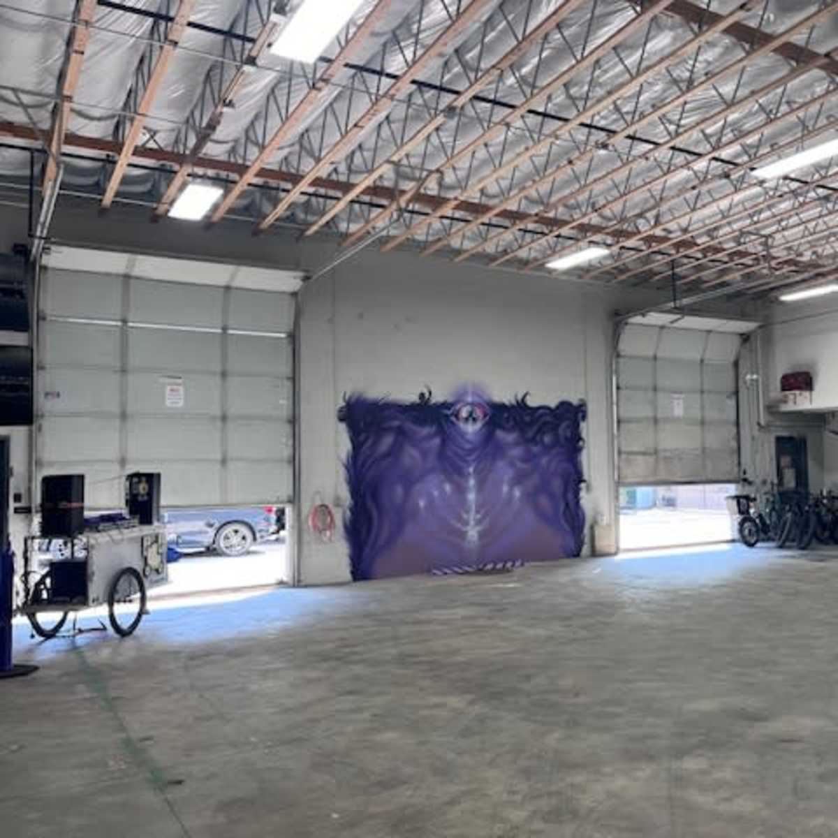 A spacious garage with a purple mural on the far wall and two large bay doors open to the outside.
