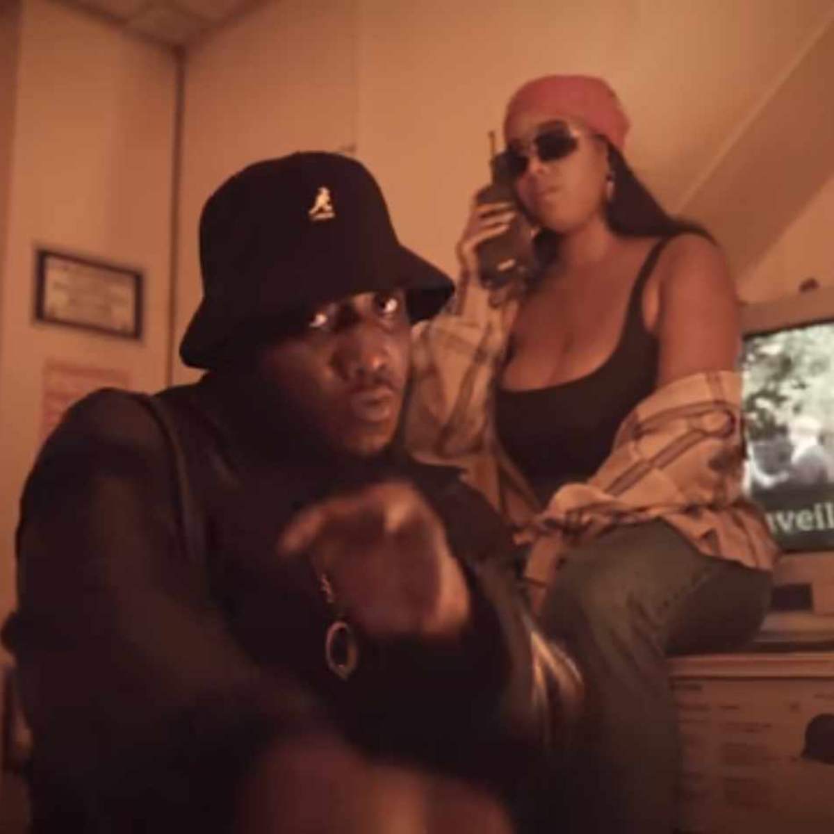 A man in a black bucket hat and sunglasses is seated in front of an old computer, while a woman wearing a pink beanie and sunglasses sits behind him, holding a phone.