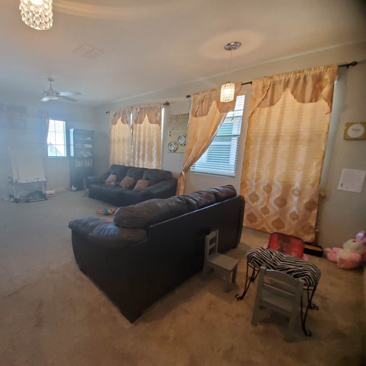 A living room with two sofas, large windows draped with sheer curtains, and a carpeted floor.