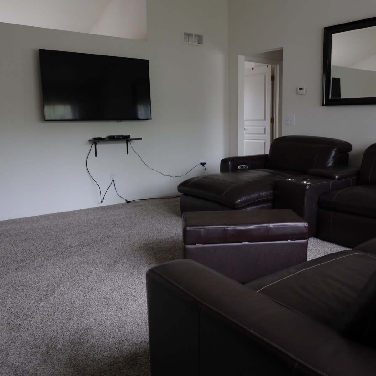 A living room with a brown sectional sofa, a large flat-screen TV mounted on the wall, and a carpeted floor.