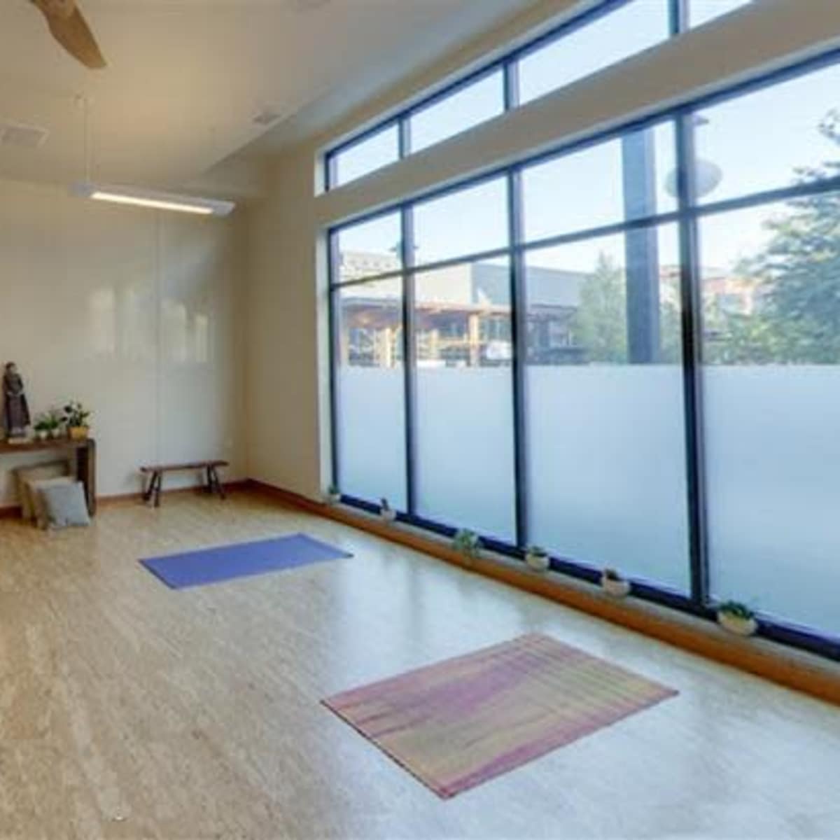 A spacious, empty studio with wooden flooring, two benches, and yoga mats placed on the ground, illuminated by natural light through large windows.