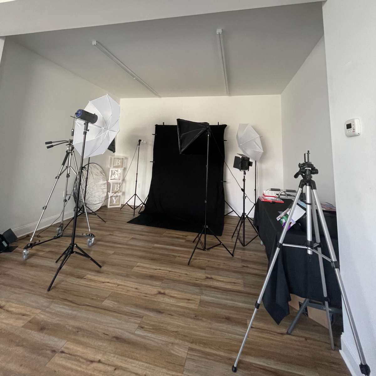 A photography studio setup with a black backdrop, lighting equipment, and tripods arranged in a spacious room with wooden flooring.