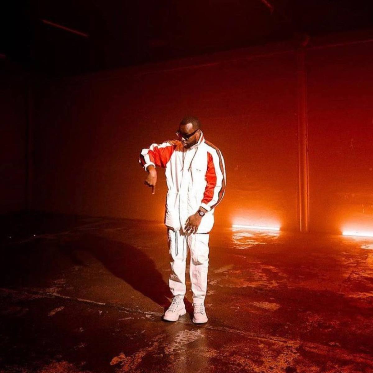 A man dressed in a white tracksuit with red sleeves stands in a dimly lit, empty warehouse space.