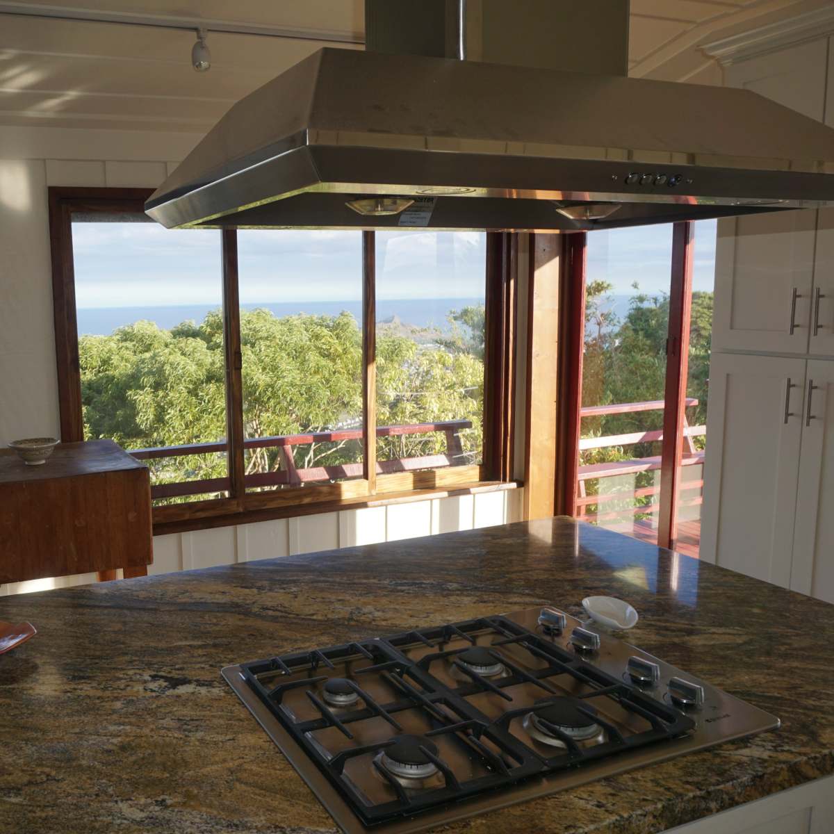 A kitchen with a gas stove on a granite countertop and large windows that offer a view of the ocean and surrounding hills.