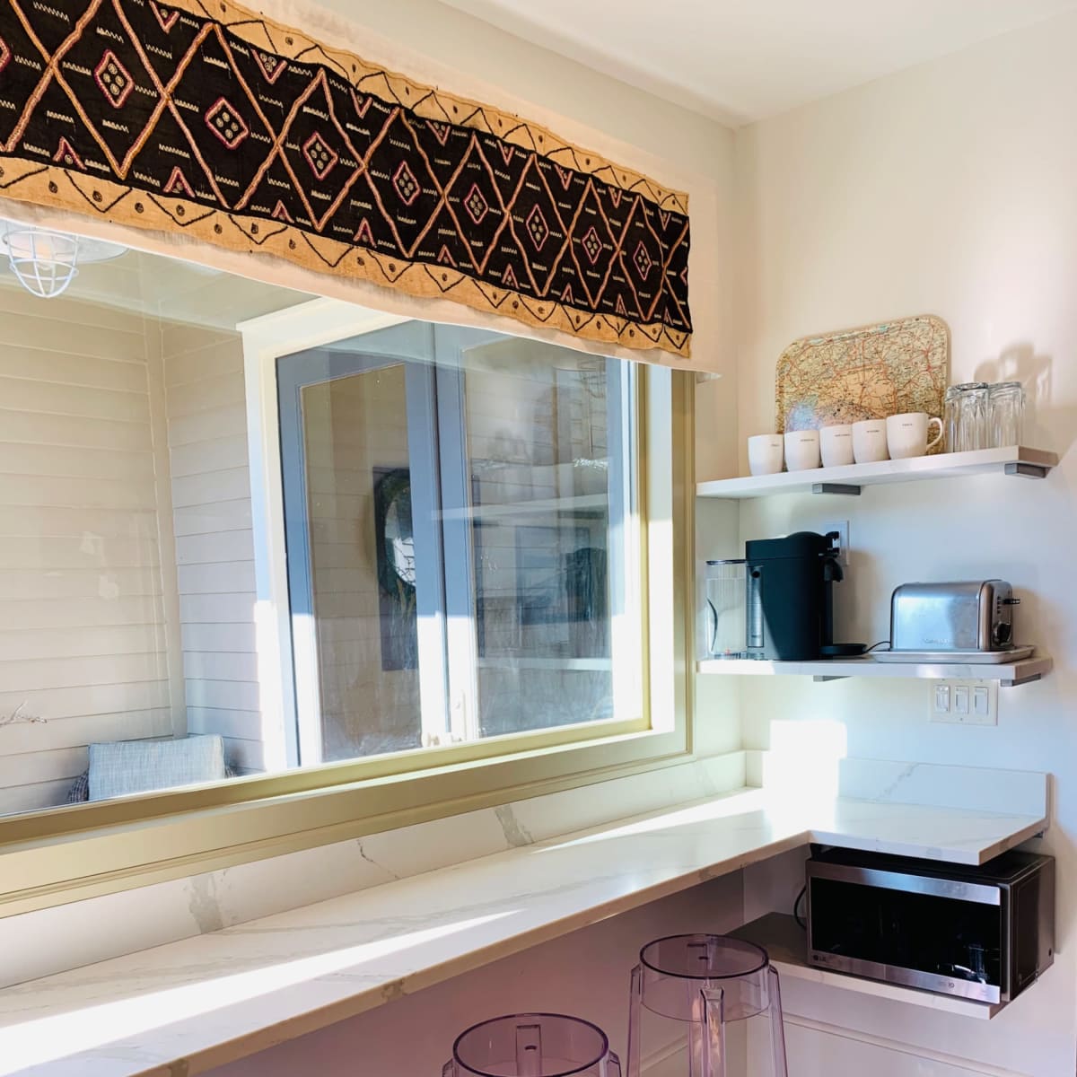 A modern kitchen area with a long countertop, three transparent stools, and shelves displaying various kitchen items.