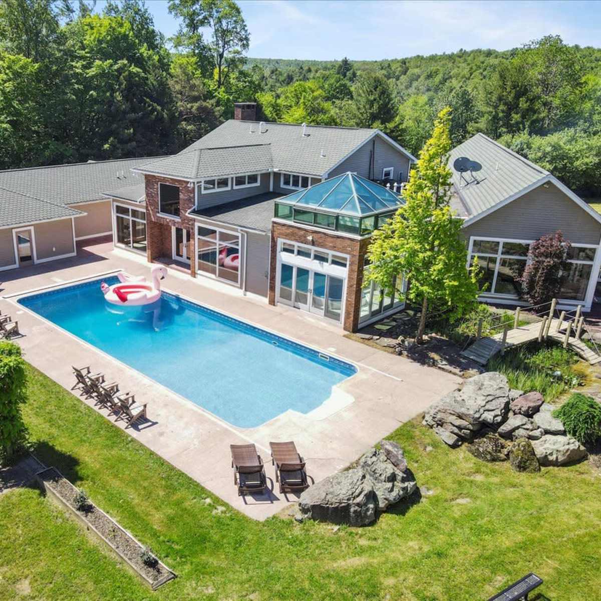 A modern house with large windows surrounds a rectangular swimming pool, featuring a giant inflatable flamingo, set against a backdrop of greenery.