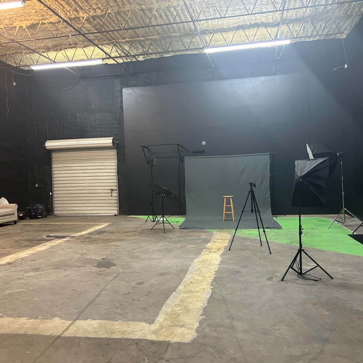 A spacious, industrial studio with black walls, various lighting equipment, and a green backdrop set up for photography or filming.