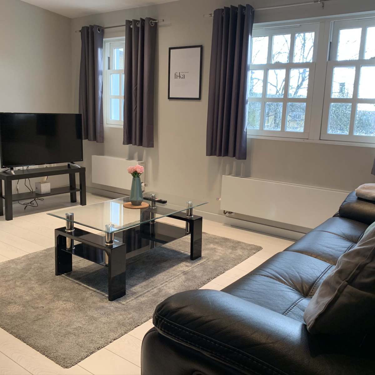 The room features a dark leather sofa, a glass coffee table on a gray rug, a television on a stand, and large windows covered with gray curtains.