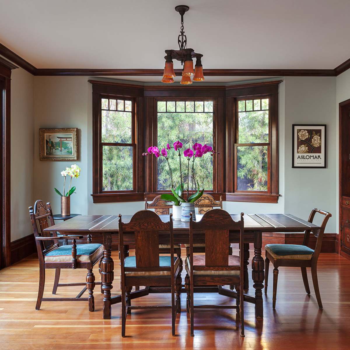 A wood-furnished dining room featuring a large table surrounded by chairs, illuminated by a pendant light and adorned with a vase of orchids.