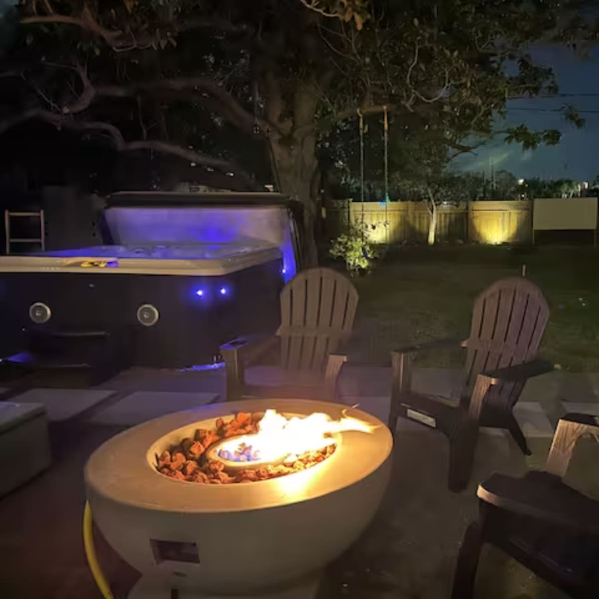 A circular fire pit surrounded by four adirondack chairs is lit in a backyard at night, with a hot tub and trees in the background.