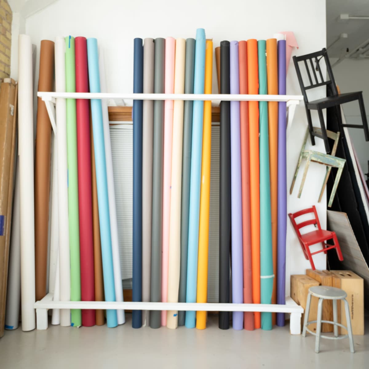 A collection of rolled-up backdrops in various colors is neatly arranged on a white rack against a wall, with a few wooden chairs and boxes nearby.