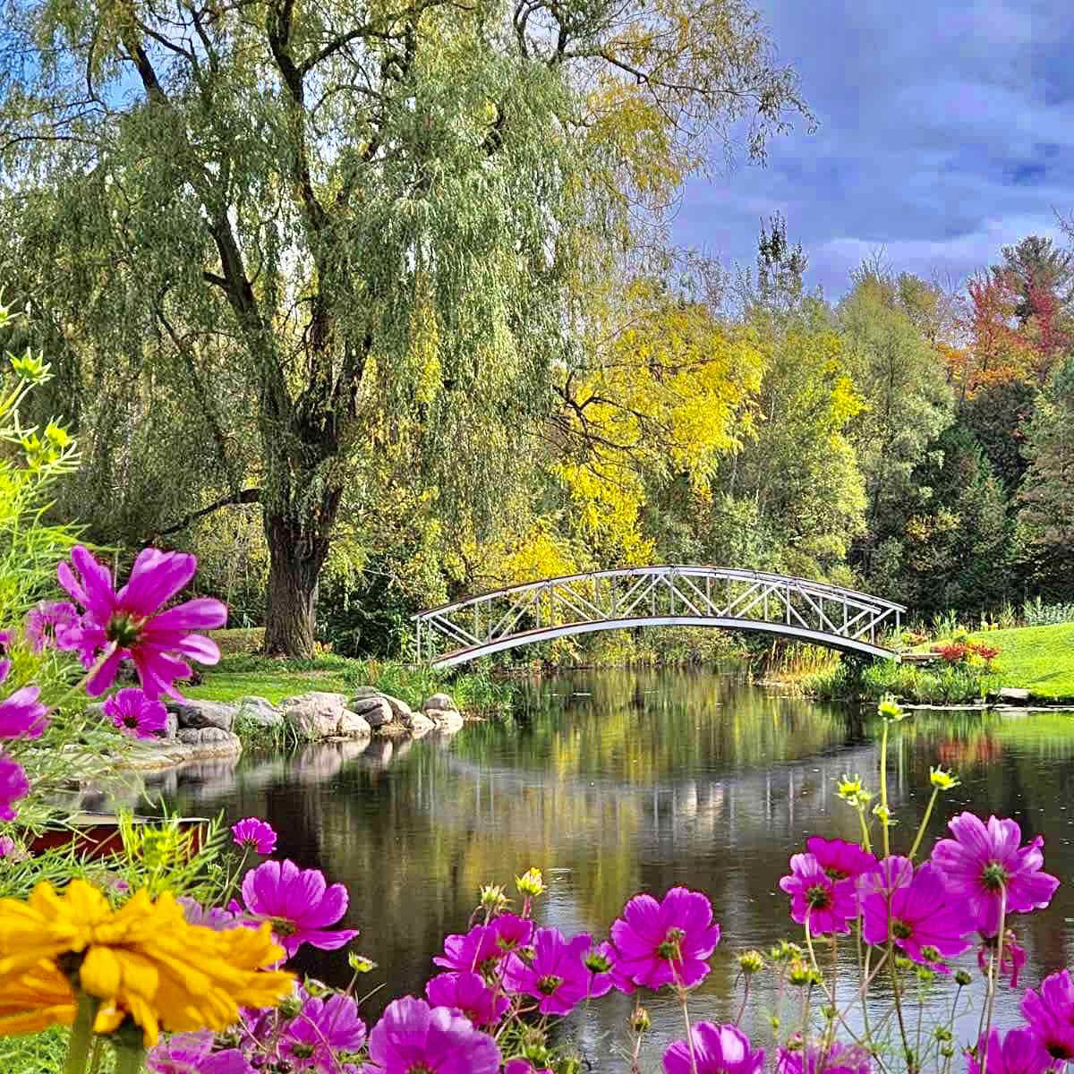A white bridge spans a calm river surrounded by trees and vibrant flowers.