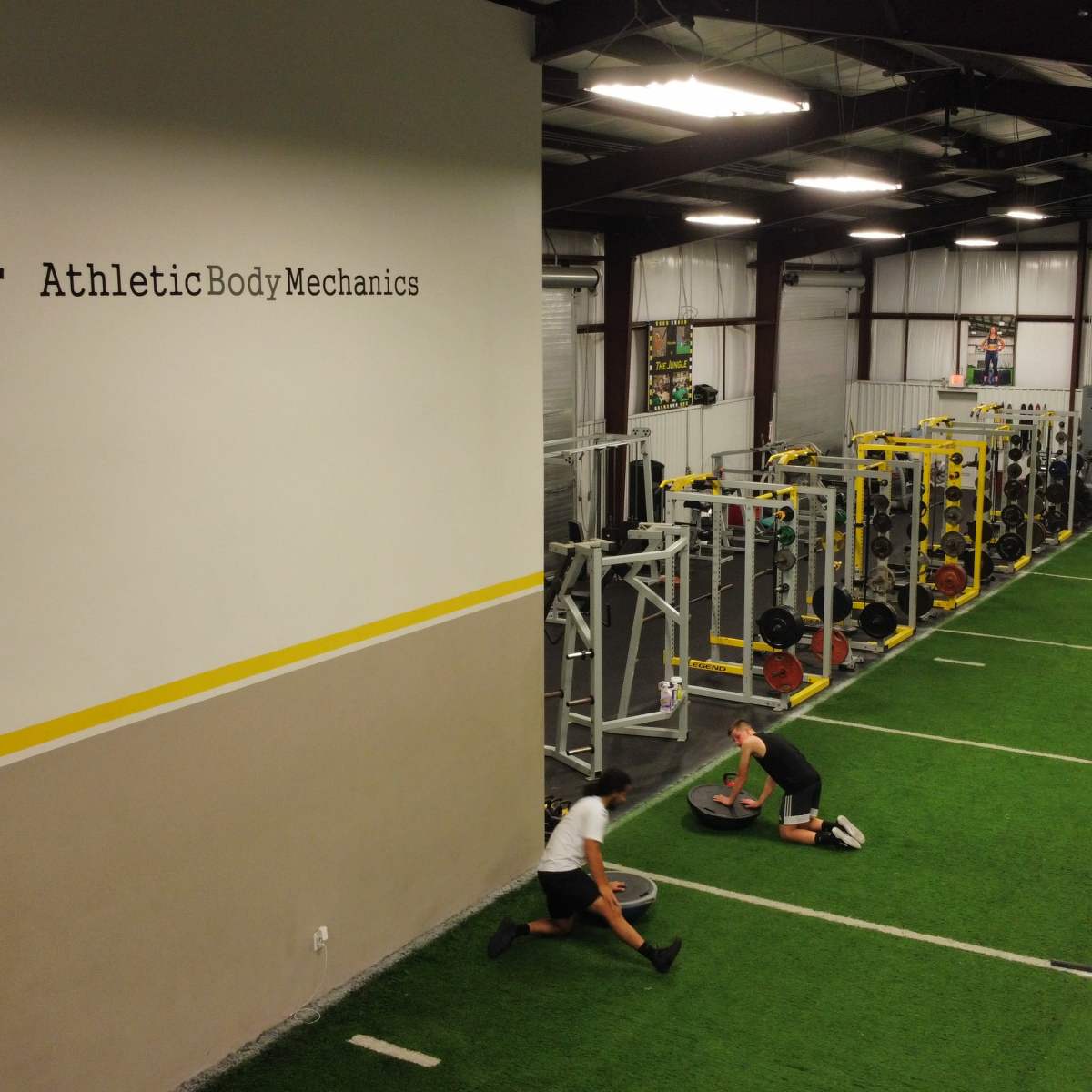 An indoor gym facility with turf flooring, equippment for strength training, and individuals engaged in fitness activities.