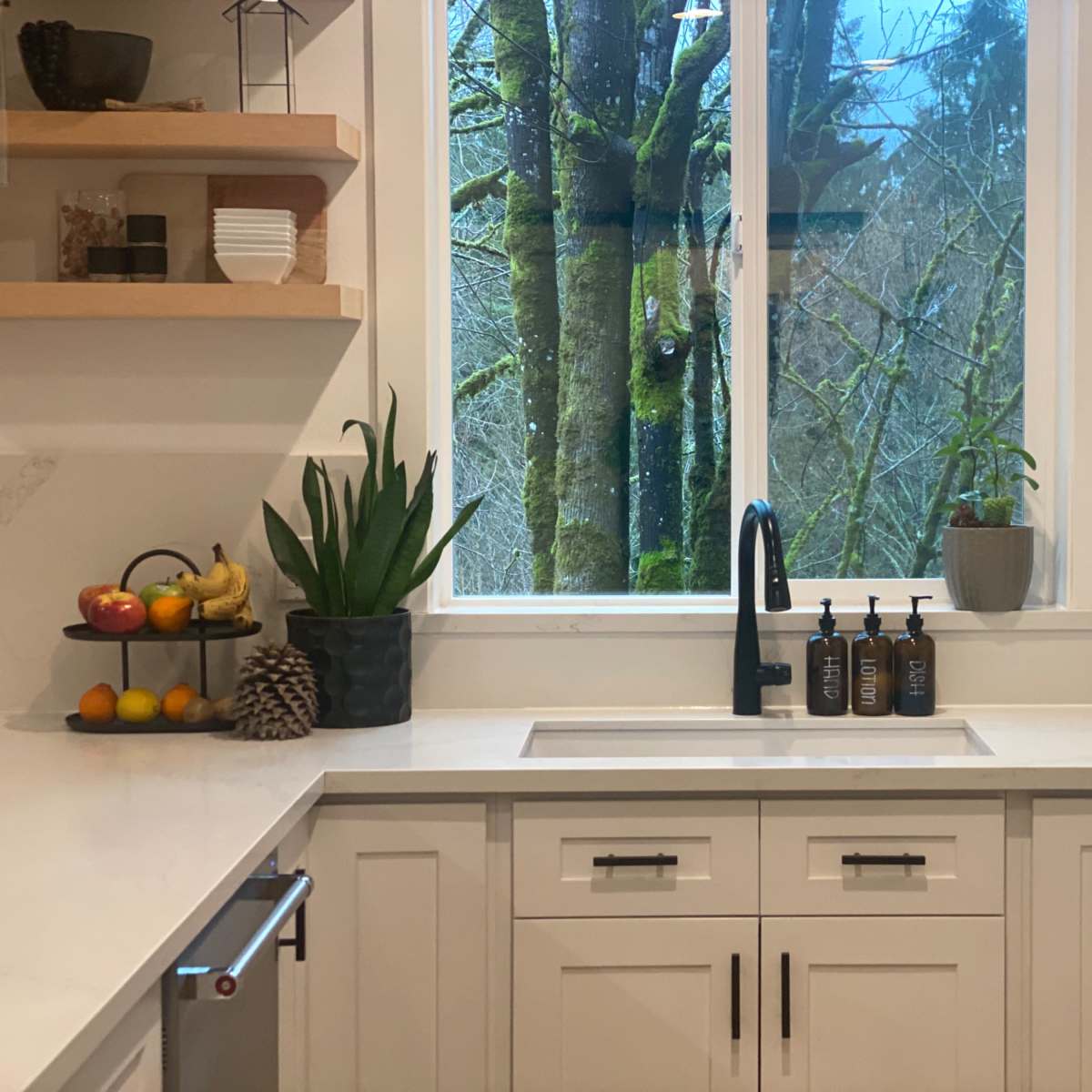 A modern kitchen features a sink beneath a large window with a view of trees, alongside minimalist cabinetry and counter arrangements.