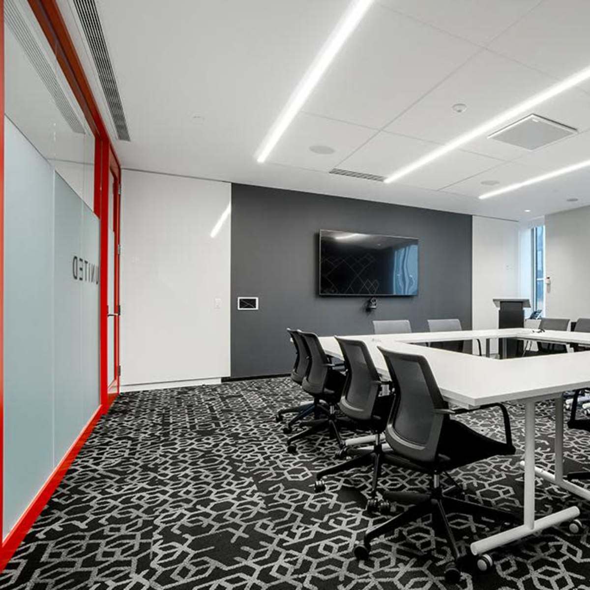 A modern conference room featuring a large rectangular table surrounded by chairs, with a television mounted on the wall and large windows providing natural light.