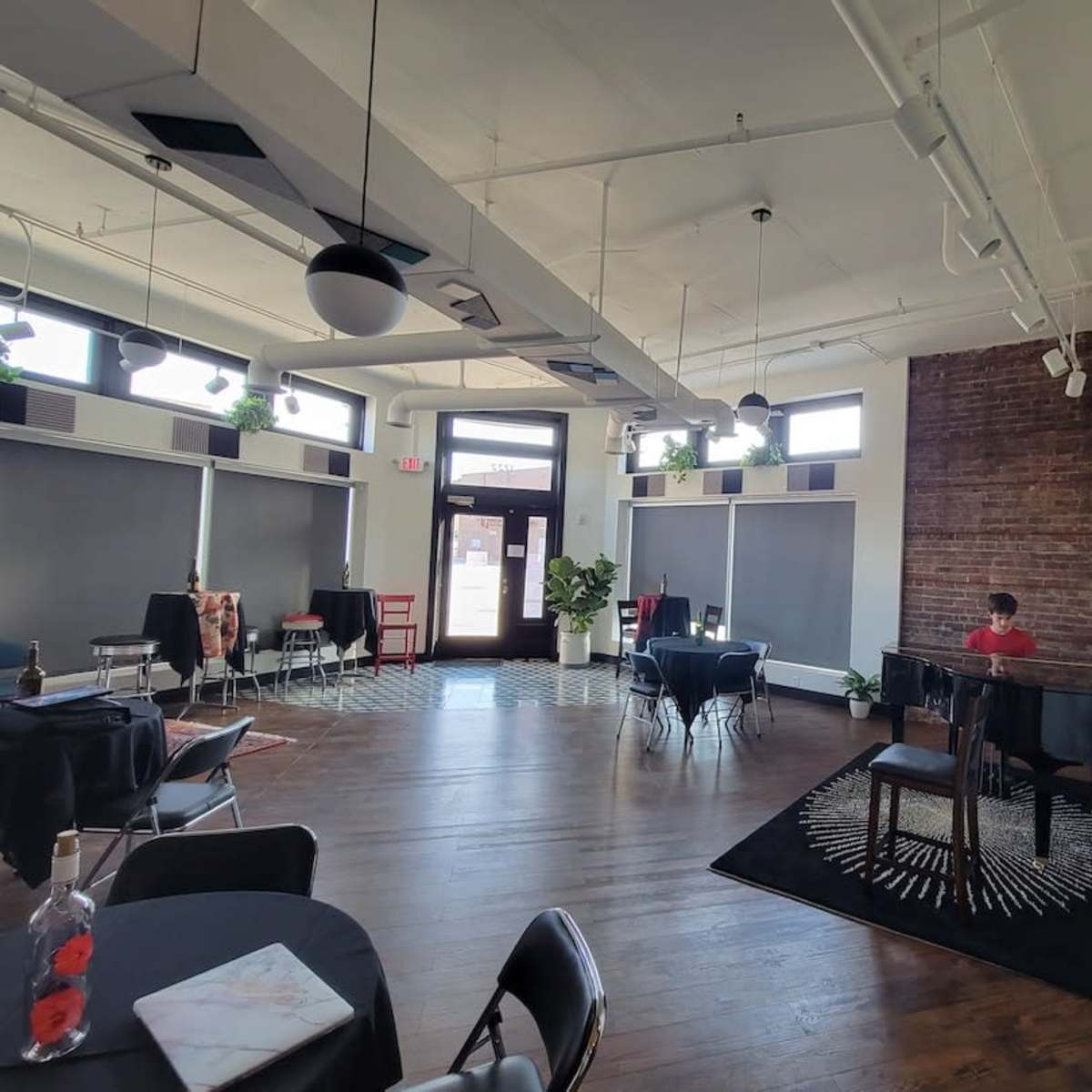 A spacious interior features wooden floors, tables set for dining, and a person seated at a piano against a backdrop of exposed brick.
