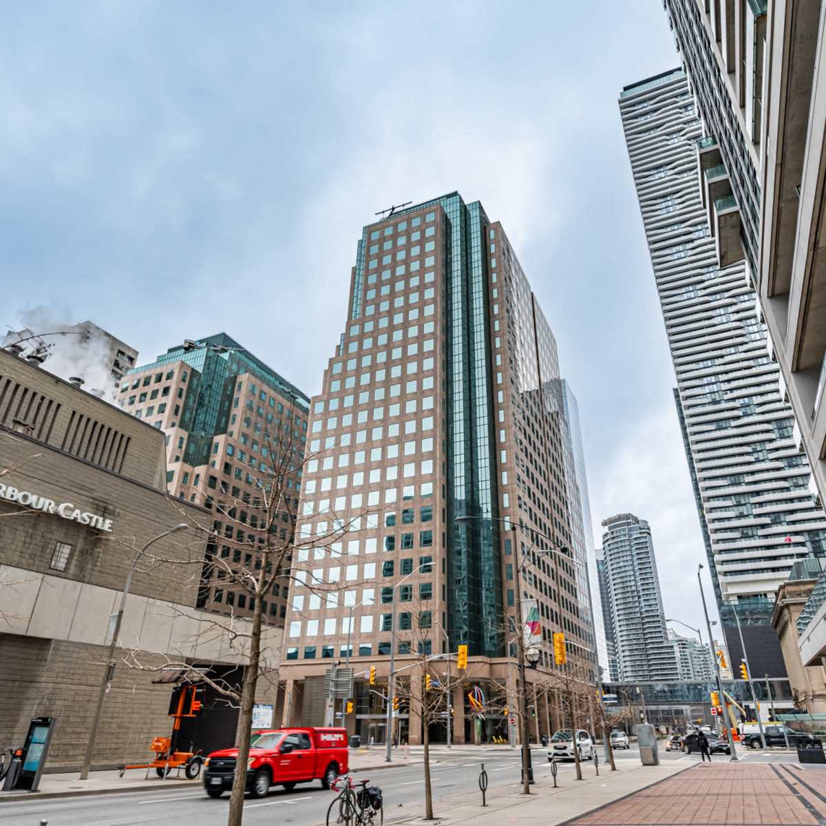 A city street lined with modern skyscrapers, featuring a mix of glass and concrete buildings under a cloudy sky.