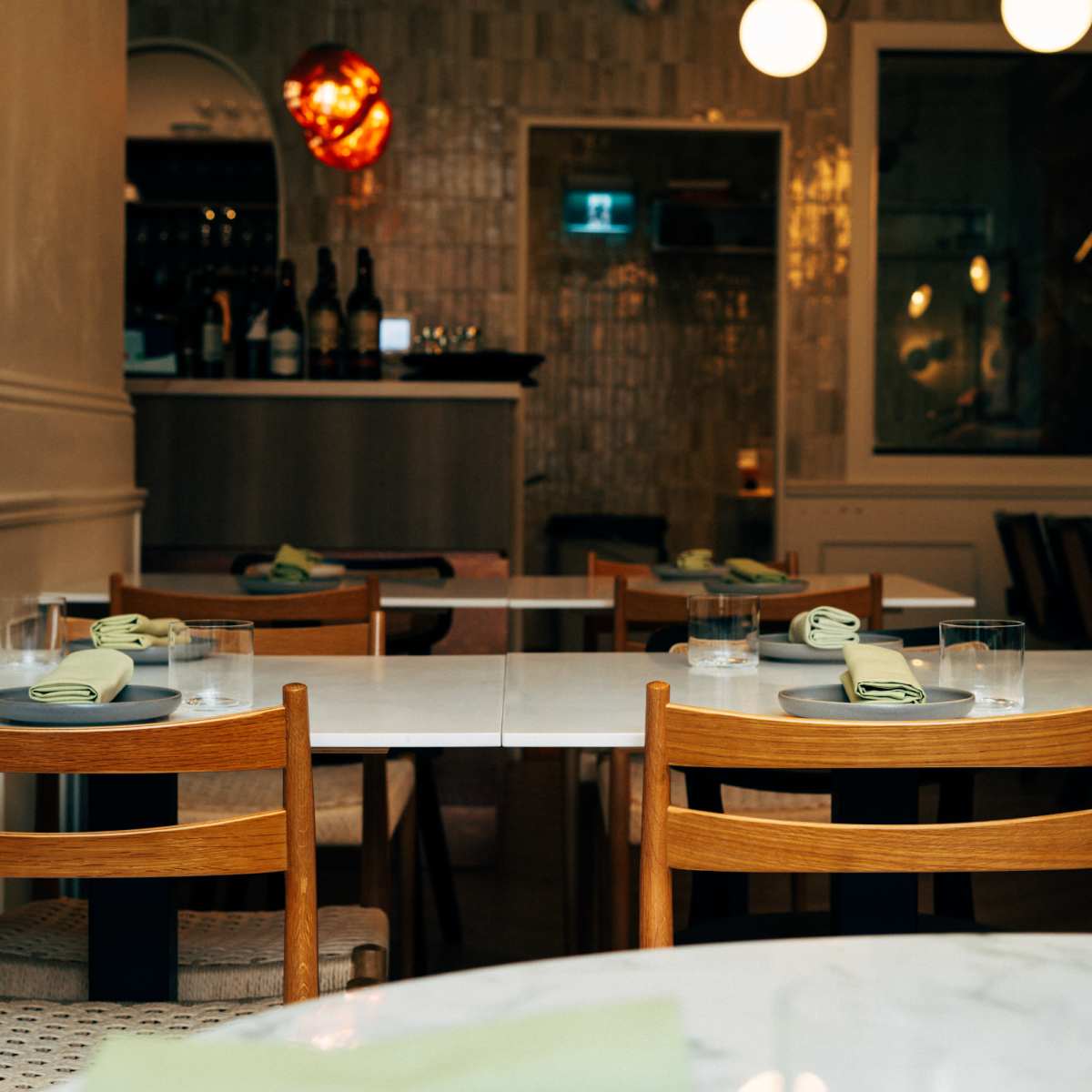 A restaurant interior features empty tables set with plates and glasses, with a bar visible in the background.
