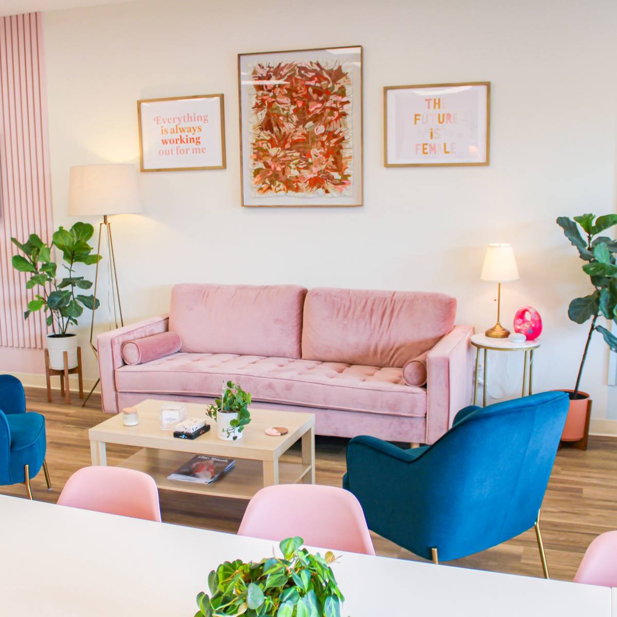 A modern, stylish living space with a pink sofa, blue chairs, a wooden coffee table, and various plants.