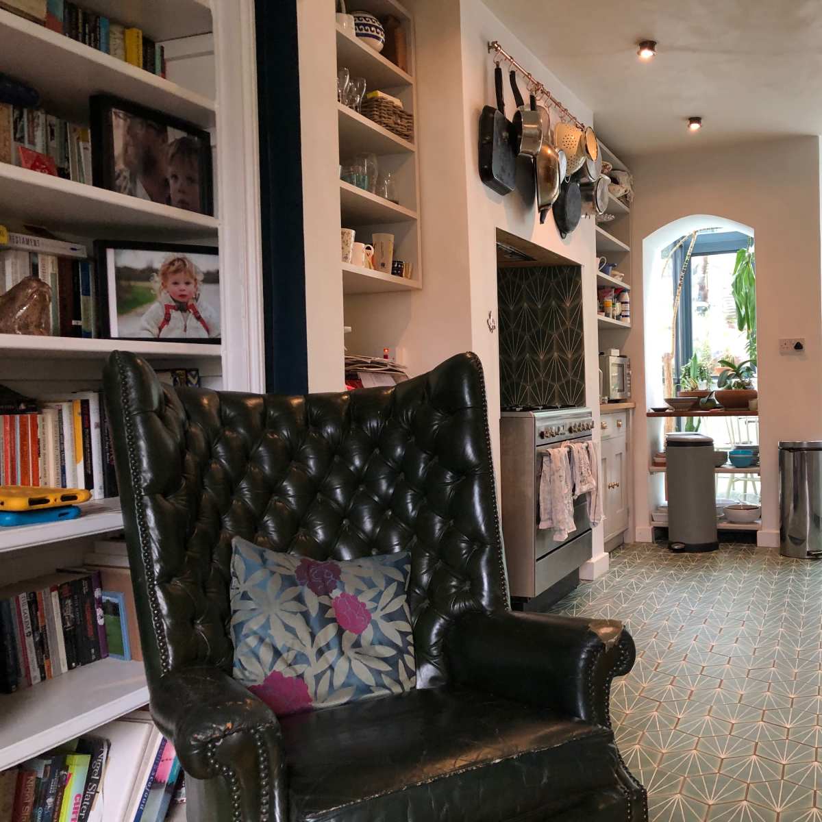 A cozy kitchen area featuring a green upholstered armchair, shelves filled with books and photos, and a view of a backyard through large windows.