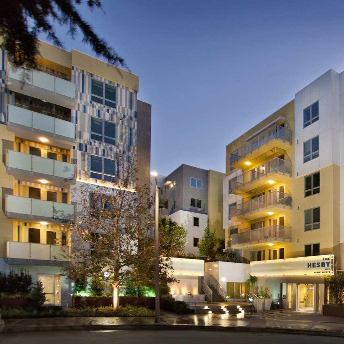 A modern apartment building with multiple stories, featuring balconies and illuminated exteriors at dusk.