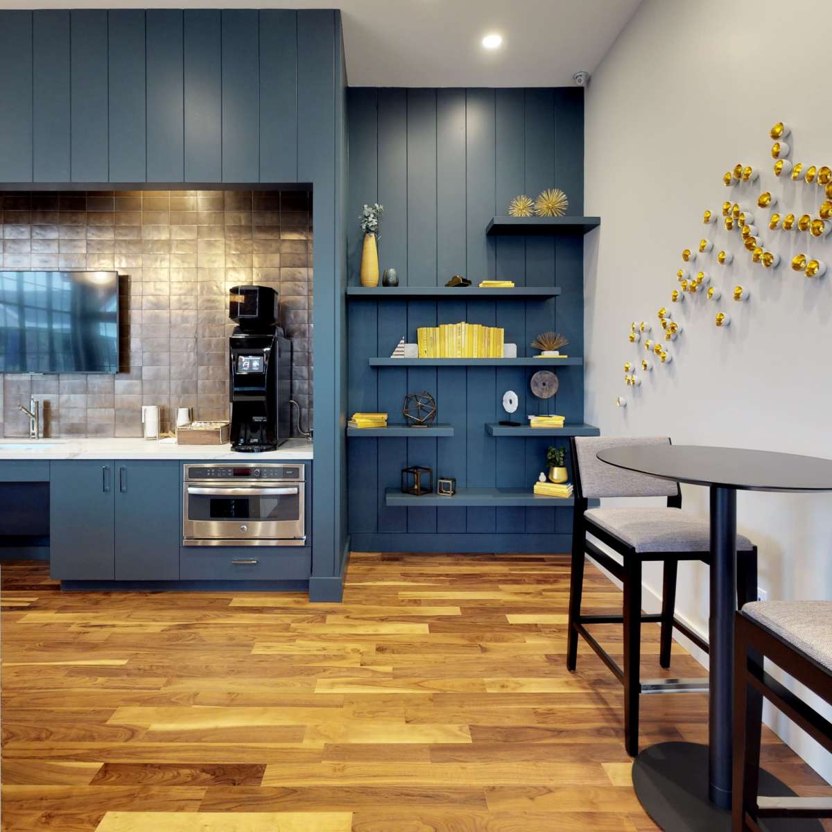 A modern kitchen area with blue cabinetry, a silver backsplash, and a small table with two chairs against a wall decorated with yellow circular accents.