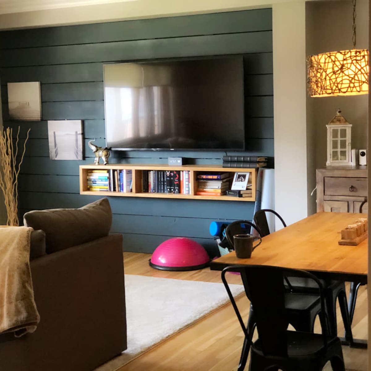 A modern living room features a large wall-mounted TV, a wooden shelf with books, and a dining table surrounded by black chairs.