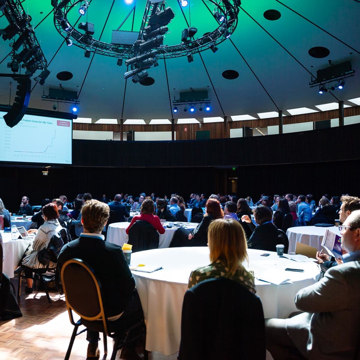 A speaker presents to an audience in a large, round room with circular lighting fixtures and a screen displaying a graph.