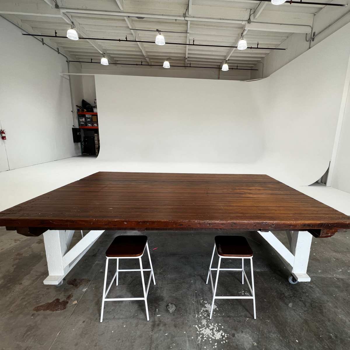 A large wooden table with two white stools is positioned in an empty studio space with a white backdrop.