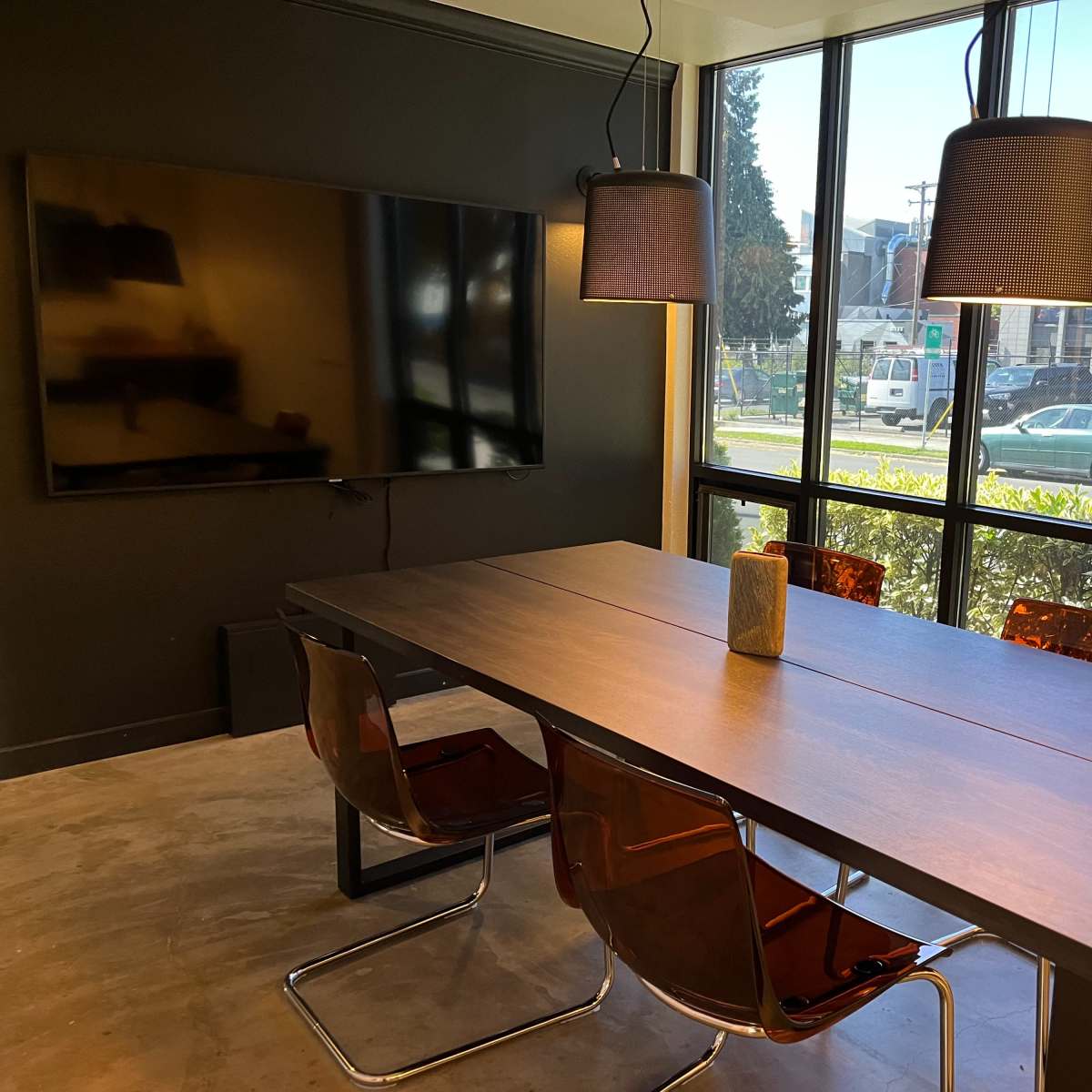 A modern conference room features a long table with four chairs, a television mounted on the wall, and large windows overlooking an outdoor area.