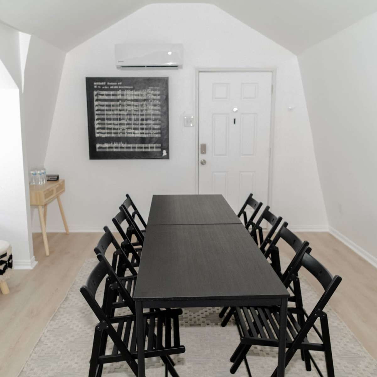 A minimalistic meeting room with a long black table surrounded by eight folding chairs, a wall-mounted air conditioner, and a large blackboard on the wall.