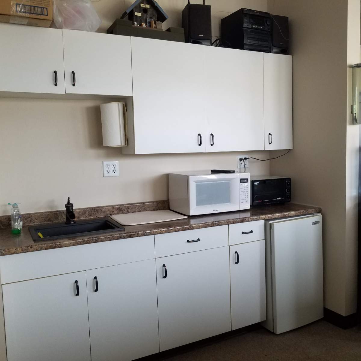 A kitchenette with white cabinets, a sink, a microwave, a small refrigerator, and a water cooler.