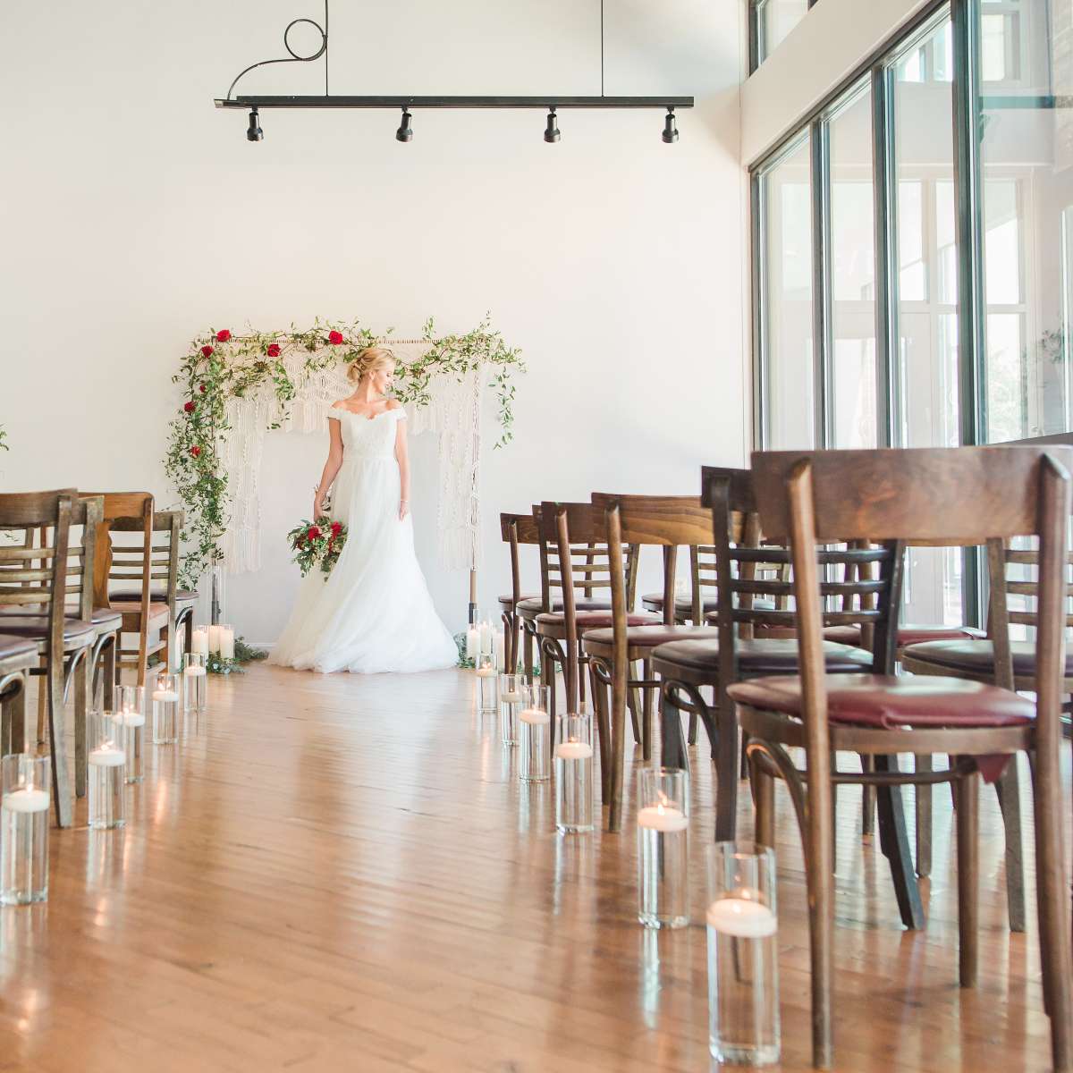 A bride stands in front of a floral archway at the end of an aisle lined with candles in a spacious, well-lit venue.