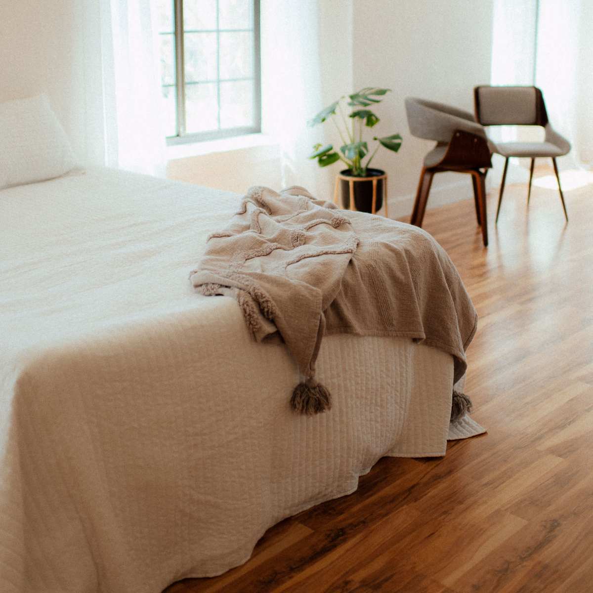 A bright bedroom features a bed with a light quilt, a plant in a pot, and a stylish chair near windows with sheer curtains.