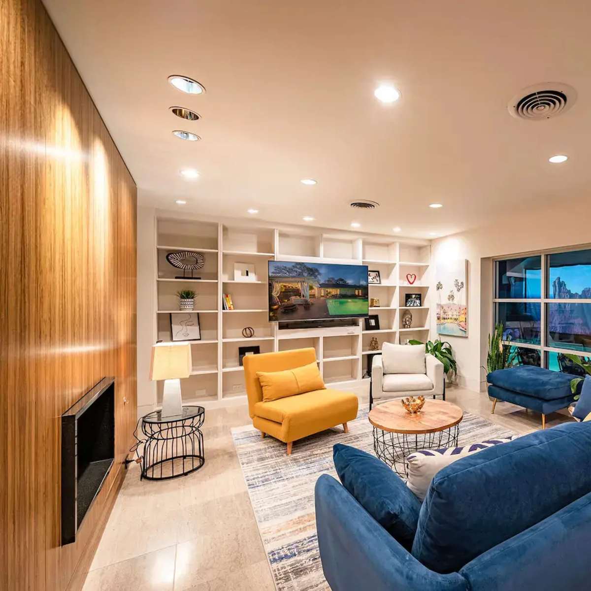 A cozy living room with a blue sofa, an orange armchair, a wooden accent wall, shelves displaying decor, and large windows overlooking a patio area.