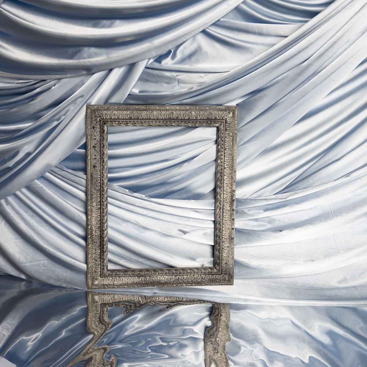 A silver ornate photo frame stands in front of a backdrop of draped blue fabric, reflecting in a smooth surface below.