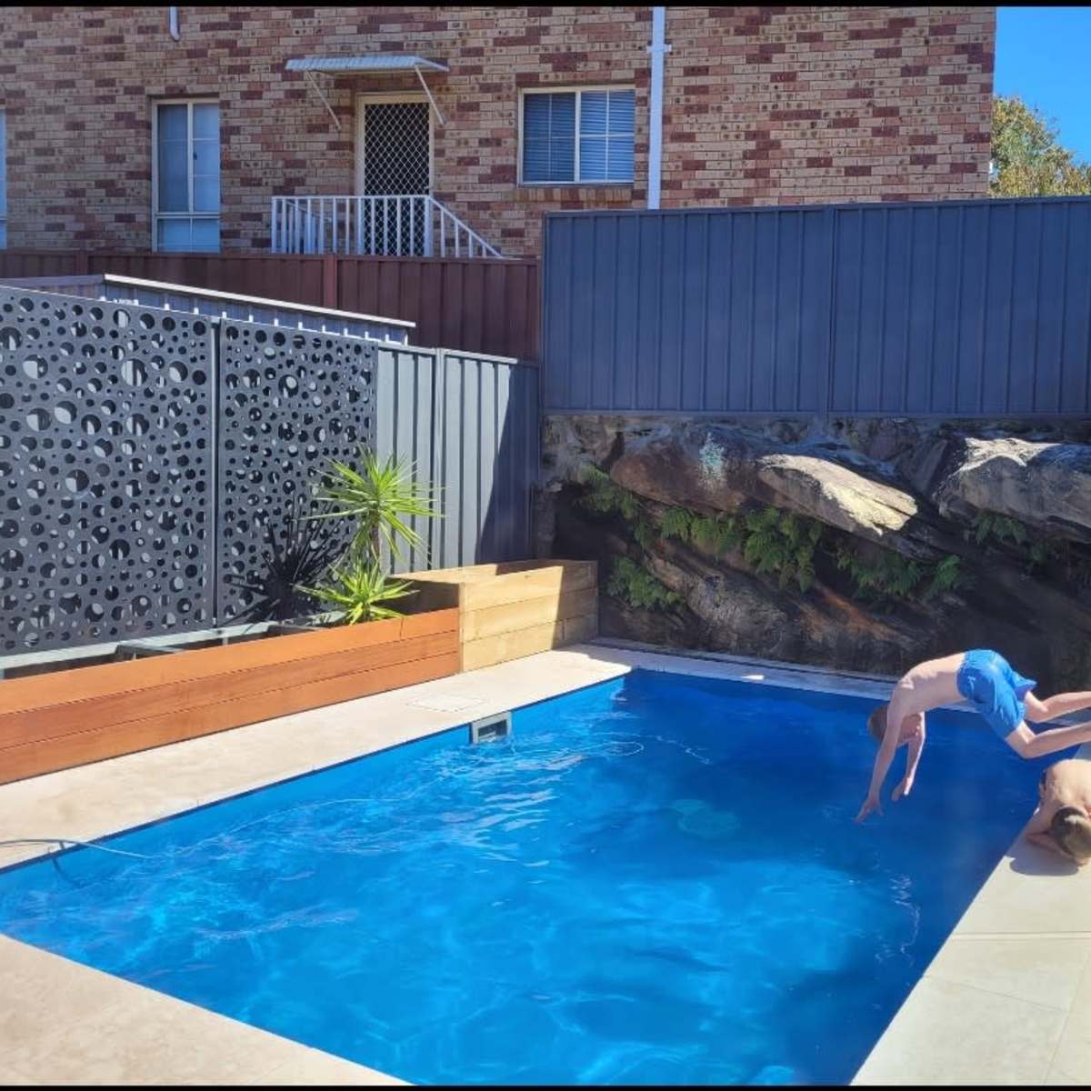 Two boys are diving into a bright blue swimming pool surrounded by a brick wall and landscaped rocks.