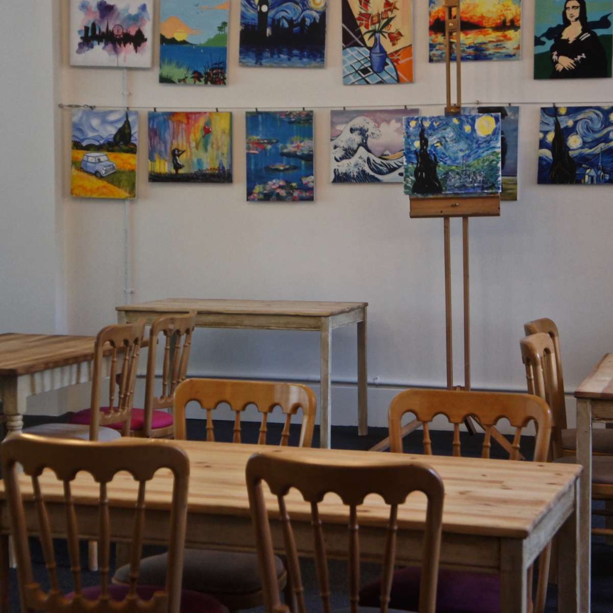 A gallery-like space with wooden tables and chairs, featuring various paintings hung on the walls.