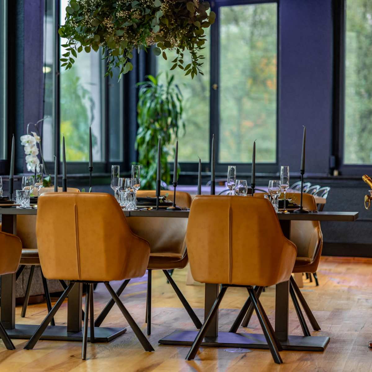 A modern dining area featuring a long table set with glassware and elegant chairs, surrounded by large windows with views of greenery outside.
