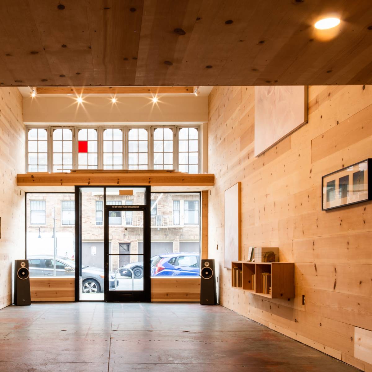 A wooden interior space with large windows, two speakers on the floor, and minimal furnishings along the walls.