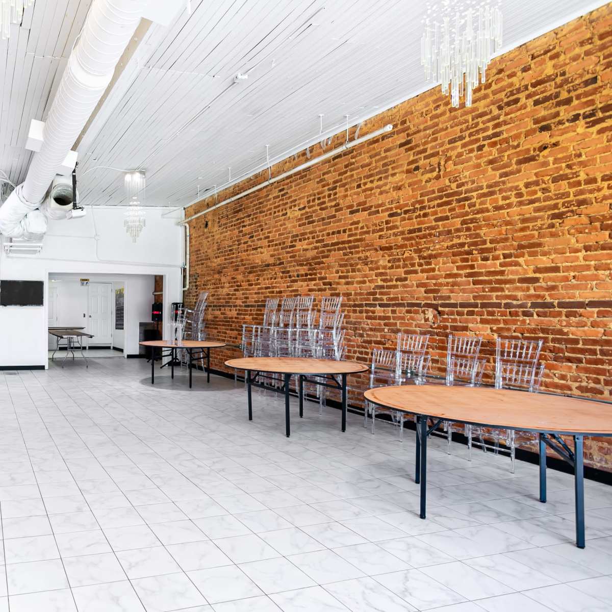 A spacious room with a white tiled floor, exposed brick walls, and several round wooden tables arranged around the space, accompanied by metal chairs stacked against the walls.