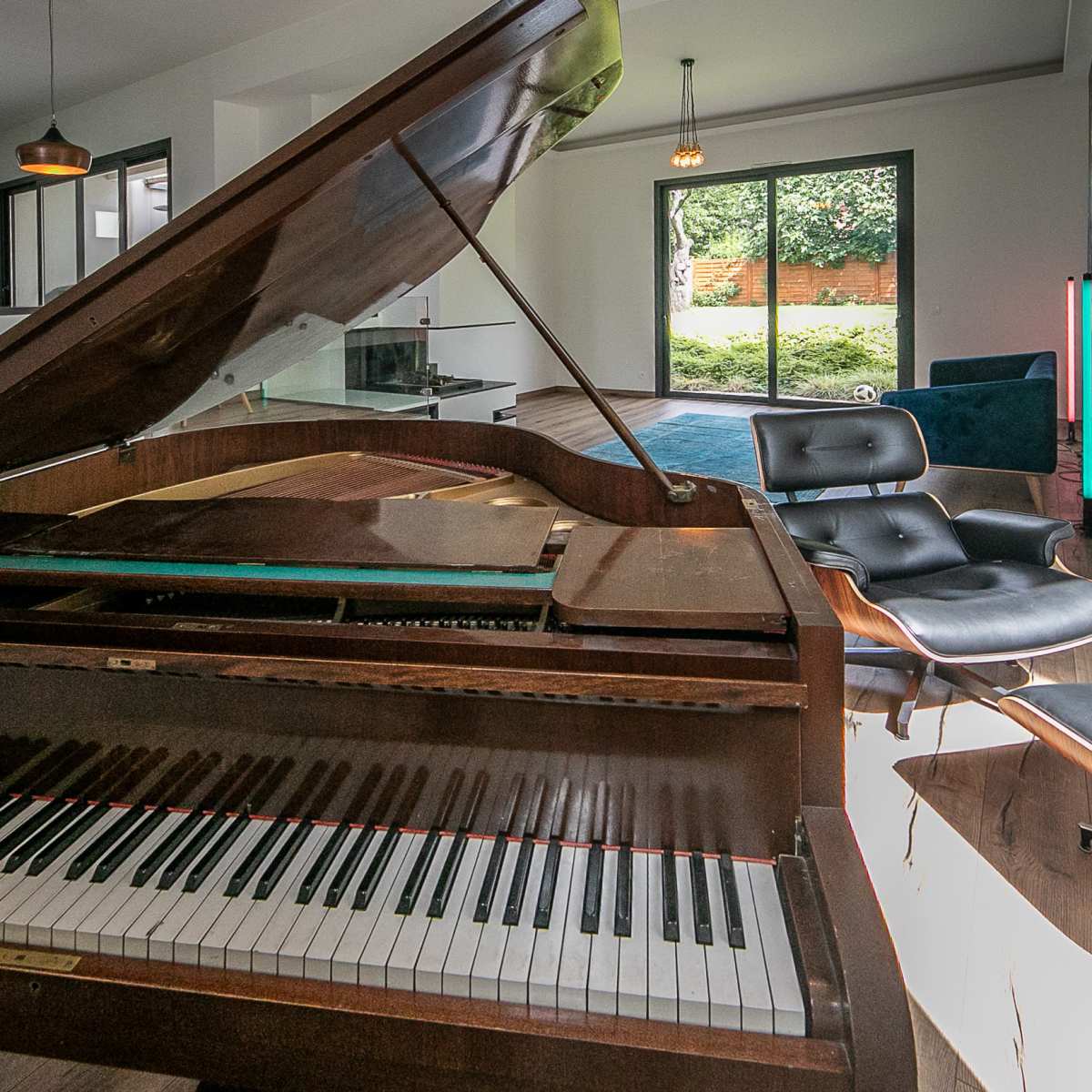 A grand piano is positioned in a modern living room, facing large glass doors that open to a landscaped backyard.