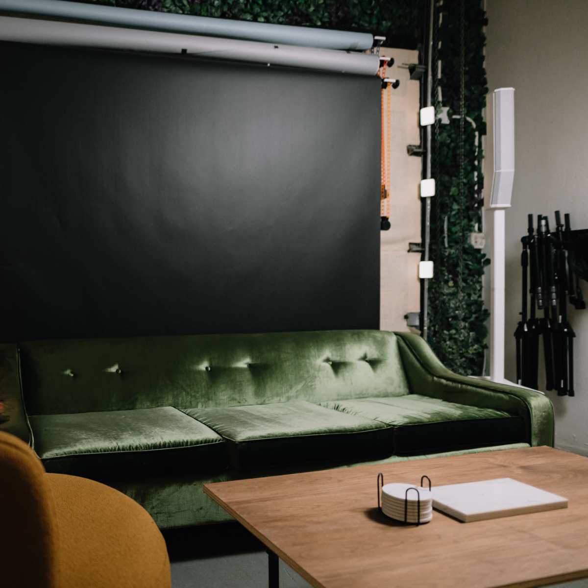 A cozy studio space featuring a green velvet sofa, an orange armchair, a large black backdrop, and plants on the walls.
