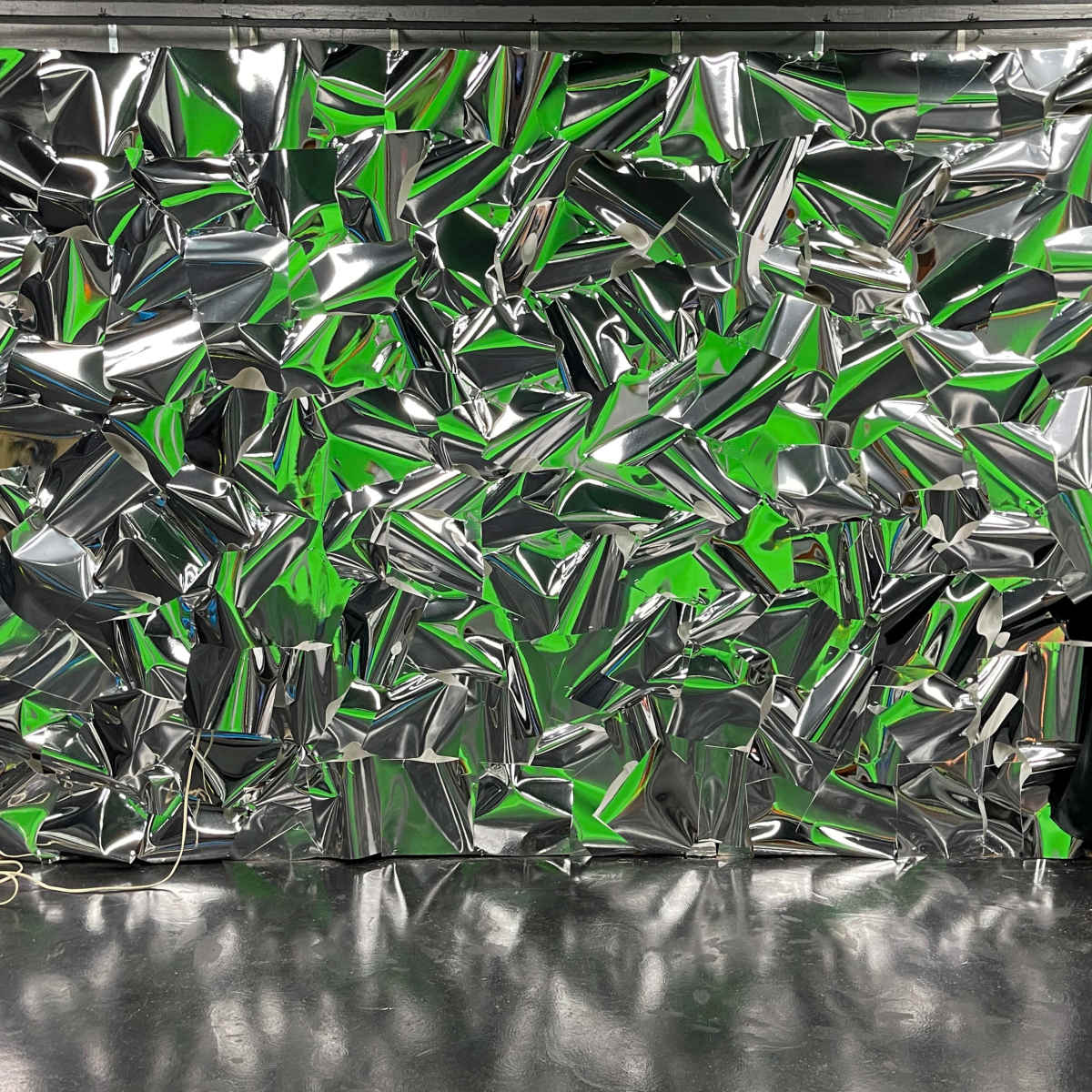 A wall covered with crumpled metallic foil in various shades of green and silver, with black curtains on either side.
